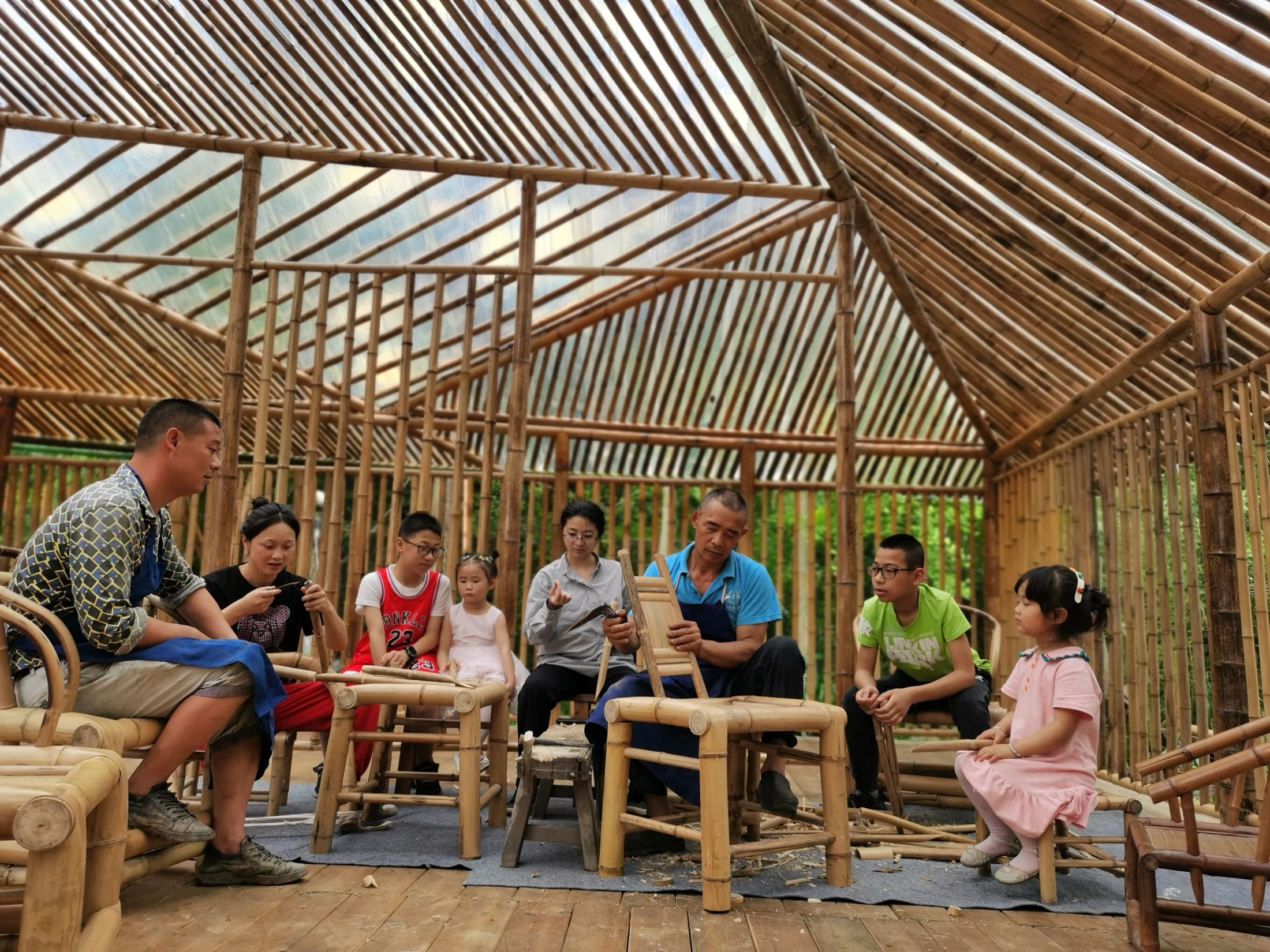 Adults and children gathered around bamboo chairs, with handwork taking place beneath the bamboo roof