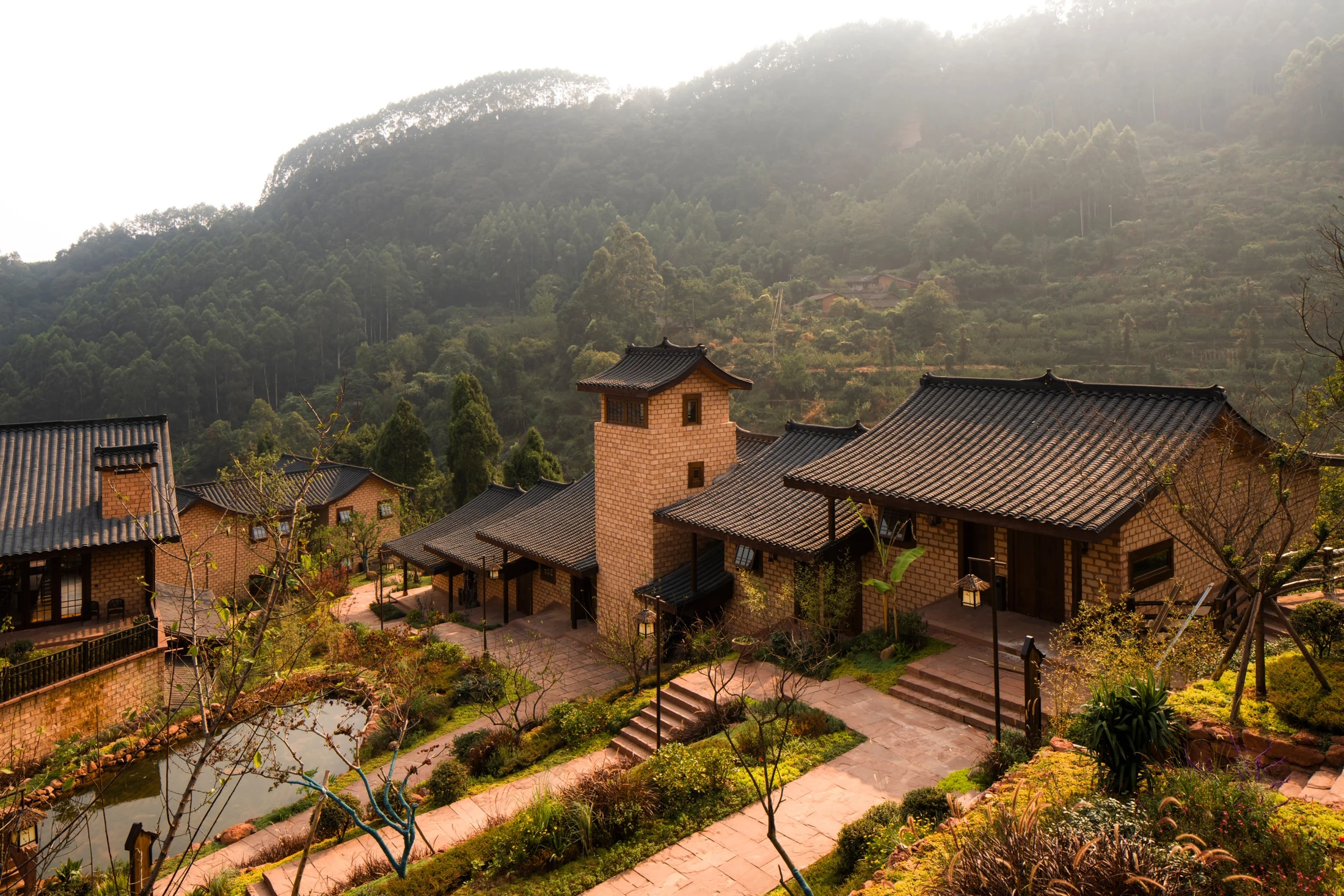 Completed buildings and courtyard paths set against a wooded hillside in warm evening light